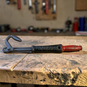 May include: A vintage Bahco nail puller tool with a black handle and a red grip. The tool is made of metal and is lying on a wooden surface. The background is a blurred workshop setting.