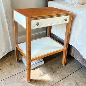 May include: A wooden bedside table with a white drawer and shelf. The top and frame are a warm brown, while the drawer and shelf have white surfaces. The drawer has a brass knob. A classic piece of bedroom furniture.
