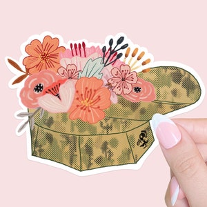 May include: A sticker featuring a floral arrangement atop a camouflage-patterned box. The flowers are in shades of peach, pink, and red, with green and brown accents. The box has a textured appearance, and the sticker is set against a pink background.