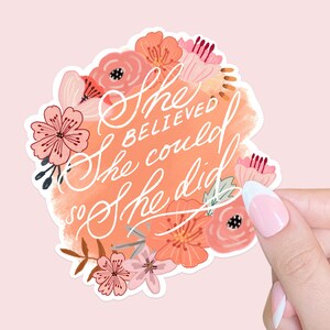 Può includere: Un adesivo floreale con la scritta "She believed she could so she did" in corsivo bianco. L'adesivo ha uno sfondo color pesca con pennellate in stile acquerello ed è circondato da fiori rosa e corallo.