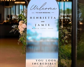 May include: A large, arched wedding sign with a white background and black text. The sign reads "Welcome to our wedding" and lists the names "Henrietta and Jamie" with the date "March 14th, 2023." The bottom of the sign says "You look incredible now let's celebrate."