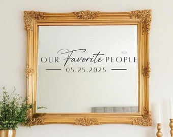 May include: A gold framed mirror with the text "OUR Favorite PEOPLE 05.25.2025". The mirror is rectangular with ornate detailing. A gold vase with greenery and two candle holders are on the mantel below.
