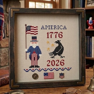May include: Framed cross-stitch artwork featuring patriotic imagery. The design includes an American flag, a figure in a top hat, and the text "AMERICA 1776 2026." The frame is dark wood with metal corner accents. Additional details include stars, a raven, and a small flag.