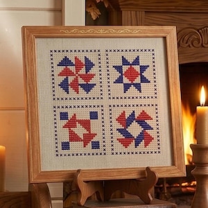 May include: A framed cross-stitch sampler featuring four geometric patterns in red and blue on a cream background. The wooden frame is displayed on a wooden easel, with a warm fireplace and candle in the background.