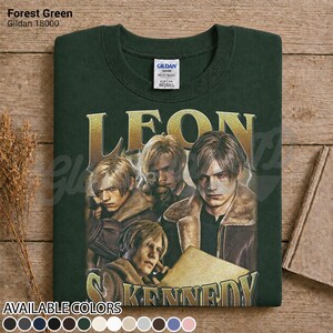 May include: A forest green Gildan 18000 sweatshirt featuring a graphic print of Leon Kennedy from Resident Evil. The design includes three depictions of the character in a brown jacket, with the name LEON KENNEDY in large gold lettering.