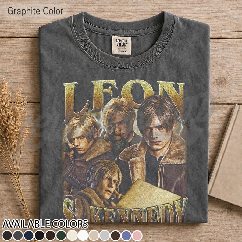 May include: A graphite-colored crew neck t-shirt featuring a graphic print of the character Leon Kennedy in multiple poses. The text LEON KENNEDY is printed in a large, gold-toned font above and below the character illustrations on the front.