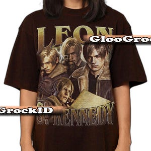 May include: A dark brown short-sleeve t-shirt featuring a graphic print of the character Leon S. Kennedy in various poses. The text LEON is printed in large gold letters at the top, and S. KENNEDY is printed in gold letters at the bottom of the graphic.