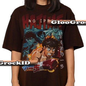 Op de afbeelding: Een donkerbruin t-shirt met korte mouwen en een grafische print van anime-personages in boksuitrusting. De tekst "HAJIME NO IPPO" staat bovenaan in het rood, met Japanse tekens en "THE FIGHTING!" onderaan het ontwerp gedrukt.