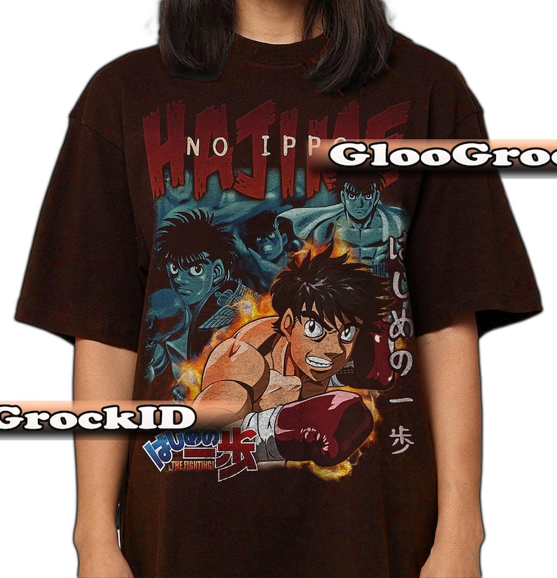 Op de afbeelding: Een donkerbruin t-shirt met korte mouwen en een grafische print van anime-personages in boksuitrusting. De tekst "HAJIME NO IPPO" staat bovenaan in het rood, met Japanse tekens en "THE FIGHTING!" onderaan het ontwerp gedrukt.