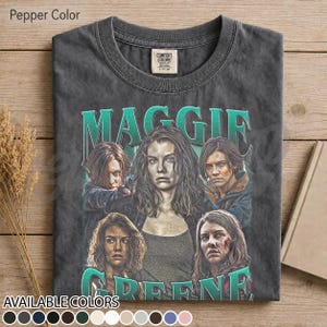 May include: A pepper-colored short-sleeve t-shirt featuring a graphic print of the character Maggie from The Walking Dead. The design includes the name MAGGIE in large teal letters above four distinct portraits of the character in various poses.