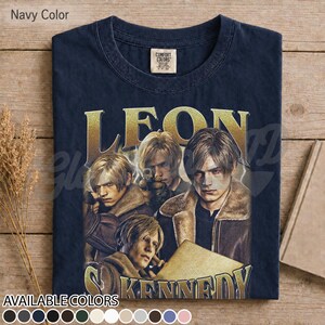 May include: A navy blue t-shirt featuring a graphic print of the character Leon Kennedy from Resident Evil. The design includes multiple depictions of the character in a brown jacket, with the name LEON KENNEDY printed in large gold letters.