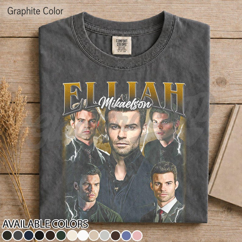 May include: Graphite grey t-shirt with a graphic of a man, the name "Elijah Mikaelson" in gold lettering, and multiple images of the man with lightning bolt accents. The shirt has a crew neck and a "Comfort Colors" tag.