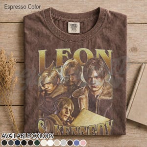 May include: An espresso-colored t-shirt featuring a graphic print of the character Leon from Resident Evil. The design includes the name LEON in large gold letters above three depictions of the character wearing a brown jacket with a fur collar.