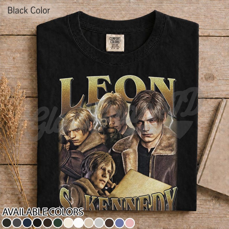 May include: A black t-shirt featuring a graphic print of the character Leon Kennedy in various poses. The text "LEON" appears in large gold letters above the character, and "KENNEDY" is printed in gold below. The shirt has a crew neckline.