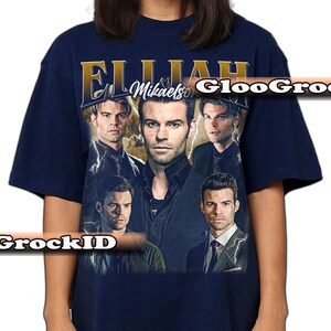 May include: Navy blue t-shirt with a collage of images of a person and the text "Elijah Mikaelson" in gold. The design includes lightning bolt graphics. A vintage-style graphic tee.