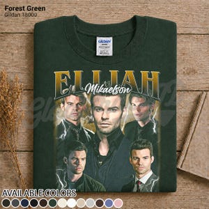 May include: Forest green Gildan 18000 sweatshirt with a gold graphic of "Elijah Mikaelson" and multiple images of a man. The sweatshirt is displayed on a wooden surface. Available in various colours.