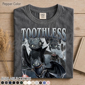 May include: A pepper-colored crew neck t-shirt featuring the text TOOTHLESS in a bold, metallic-style font. Below the text, a graphic displays multiple black dragon characters with large green eyes and wings, set against a dark, stormy background with lightning bolts.