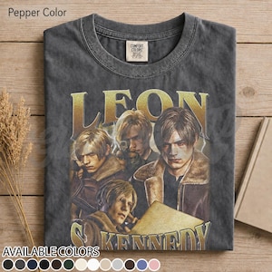 May include: A charcoal gray crew neck t-shirt featuring a graphic print of the character Leon Kennedy from Resident Evil. The design includes the name LEON KENNEDY in large gold lettering and multiple depictions of the character in a jacket.