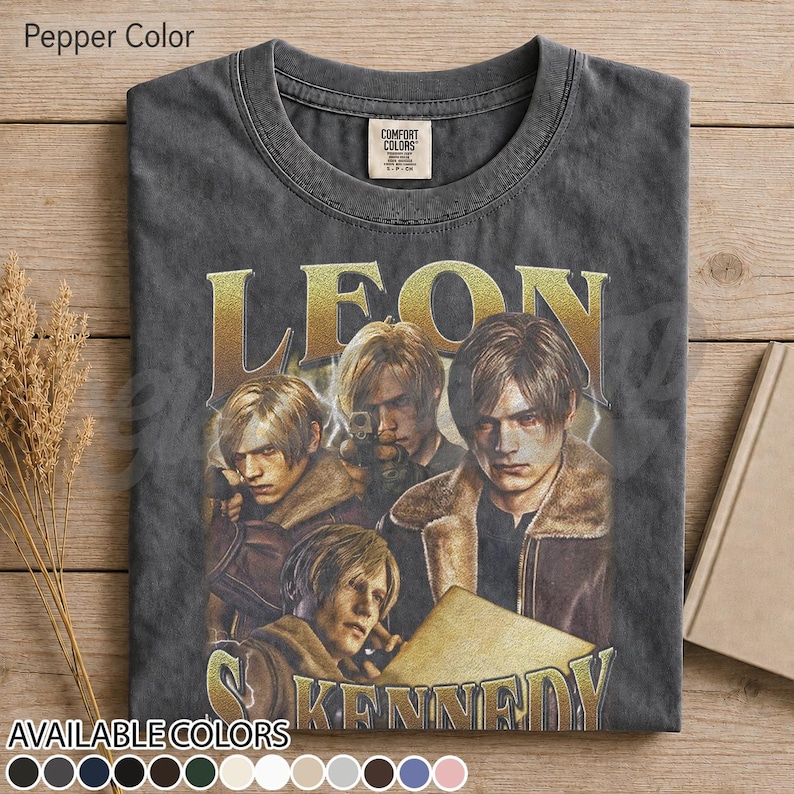 May include: A charcoal gray crew neck t-shirt featuring a graphic print of the character Leon Kennedy from Resident Evil. The design includes the name LEON KENNEDY in large gold lettering and multiple depictions of the character in a jacket.