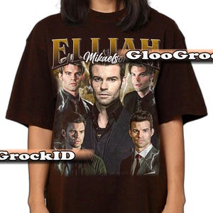 May include: A brown t-shirt with a design featuring multiple images of a man, with the name "Elijah Mikaelson" in gold. The design incorporates lightning bolt graphics and several portraits of the same person.
