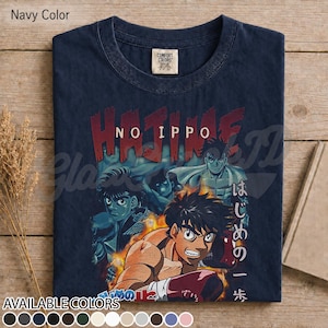 Op de afbeelding: Een marineblauw t-shirt met een grafische print van de anime Hajime no Ippo. Het ontwerp toont meerdere personages in bokshoudingen met vlammen, de tekst "HAJIME NO IPPO" in het rood en Japanse karakters verticaal aan de rechterkant.