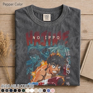 Op de afbeelding: Een peperkleurig t-shirt met ronde hals en een grafische print van de anime Hajime No Ippo. Het ontwerp toont meerdere personages in een boksthema met rode tekst Hajime No Ippo en Japanse karakters aan de rechterkant van de afbeelding.
