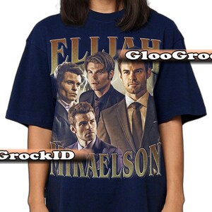 K&ouml;nnte beinhalten: Marineblaues T-Shirt mit einem Vintage-Design, das vier M&auml;nner und den goldenen Text "ELIJAH MIKAELSON" zeigt. Das Rundhals-T-Shirt ist wahrscheinlich aus Baumwolle. Das Design hat ein abgenutztes, gealtertes Aussehen.