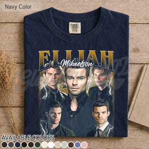 May include: Navy blue t-shirt with a graphic of Elijah Mikaelson and gold text reading "Elijah Mikaelson". The shirt has a vintage appearance and displays a range of available colours at the bottom.