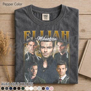 May include: Pepper-coloured t-shirt featuring a graphic of Elijah Mikaelson with multiple images of the character. The text "Elijah Mikaelson" is in gold lettering. The shirt has a crew neck and is made of a soft material. Available in multiple colours.