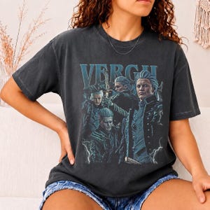 May include: A charcoal gray short-sleeve t-shirt featuring a graphic print of the character Vergil from the Devil May Cry series. The design includes the text "VERGIL" in a light blue font above multiple depictions of the character with a sword.