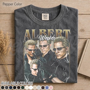 Può includere: Una t-shirt color pepe con una stampa grafica di Albert Wesker in tre pose diverse, tutti con occhiali da sole. Il testo "ALBERT" appare in grandi lettere dorate sopra il personaggio, con "Wesker" scritto in corsivo bianco.