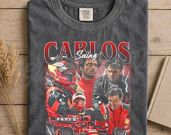 Op de afbeelding: Pepperkleurig t-shirt met een vintage afbeelding van Carlos Sainz, een Formule 1-coureur. Het ontwerp bevat afbeeldingen van Sainz in zijn race-uitrusting en de tekst "CARLOS Sainz" in het rood. Verkrijgbaar in meerdere kleuren.