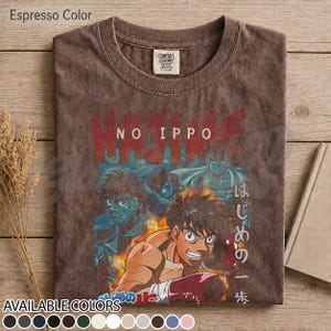 Op de afbeelding: Een espresso-kleurig t-shirt met korte mouwen en een grafische print van personages uit de anime Hajime No Ippo. Het ontwerp bevat de tekst Hajime No Ippo in het rood en Japanse karakters. Het shirt heeft een ronde hals en een Comfort Colors label.