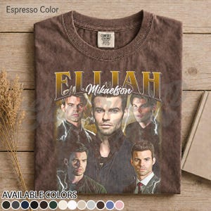 May include: Espresso-coloured t-shirt with a vintage-style graphic of a man, the name "Elijah Mikaelson" in gold lettering. The design includes multiple images of the same person, with lightning bolt accents. The shirt has a crew neck and a distressed appearance.