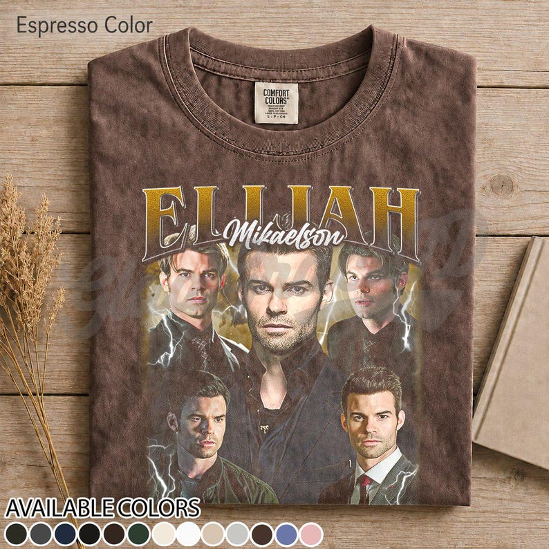 May include: Espresso-coloured t-shirt with a vintage-style graphic of a man, the name "Elijah Mikaelson" in gold lettering. The design includes multiple images of the same person, with lightning bolt accents. The shirt has a crew neck and a distressed appearance.
