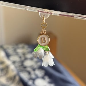 May include: A gold-colored charm with a white flower design and the letter "B" on a Kindle. The charm has a green and white accent and is attached to the Kindle with a clear plastic clip. The word "kindle" is printed on the device.