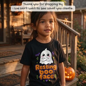 May include: A black t-shirt featuring a white cartoon ghost with a grumpy expression and crossed arms. Below the ghost, the text reads 'Resting boo face' in orange and purple letters, surrounded by small purple bats and yellow stars.