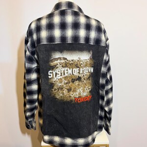 May include: A long-sleeve button-up shirt featuring a black and white plaid pattern. The back panel is a solid black fabric with a graphic print that reads System of a Down and Toxicity in white and red text over a textured landscape image.