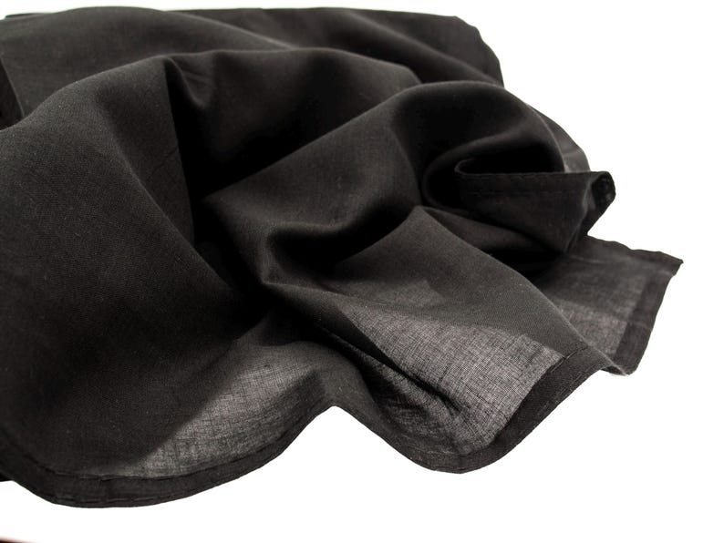 May include: A close-up of a black fabric, possibly a scarf or shawl, with soft folds and a slightly translucent appearance. The fabric is a solid black color and appears to be lightweight.