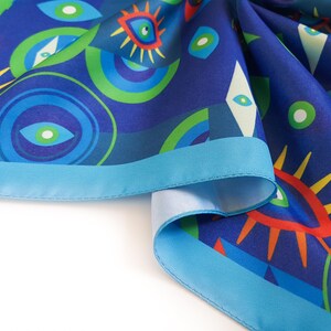 May include: A blue silk scarf with a colorful eye pattern and a turquoise border. The scarf is folded and draped on a white surface.
