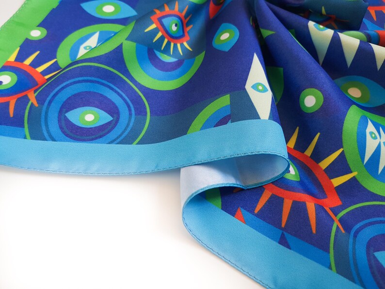 May include: A blue silk scarf with a colorful eye pattern and a turquoise border. The scarf is folded and draped on a white surface.