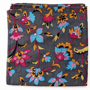May include: Folded square textile with a floral pattern. The fabric is dark gray, with a design of pink, blue, orange, and yellow flowers and leaves. The edges of the fabric are frayed.