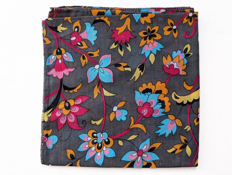 May include: Folded square textile with a floral pattern. The fabric is dark gray, with a design of pink, blue, orange, and yellow flowers and leaves. The edges of the fabric are frayed.