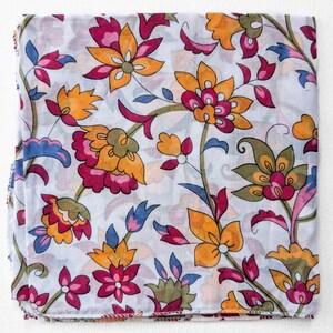 May include: A folded white fabric with a vibrant floral pattern. The design features large, stylized flowers in shades of orange, pink, and green, with blue accents. The fabric is likely a scarf or a similar textile.
