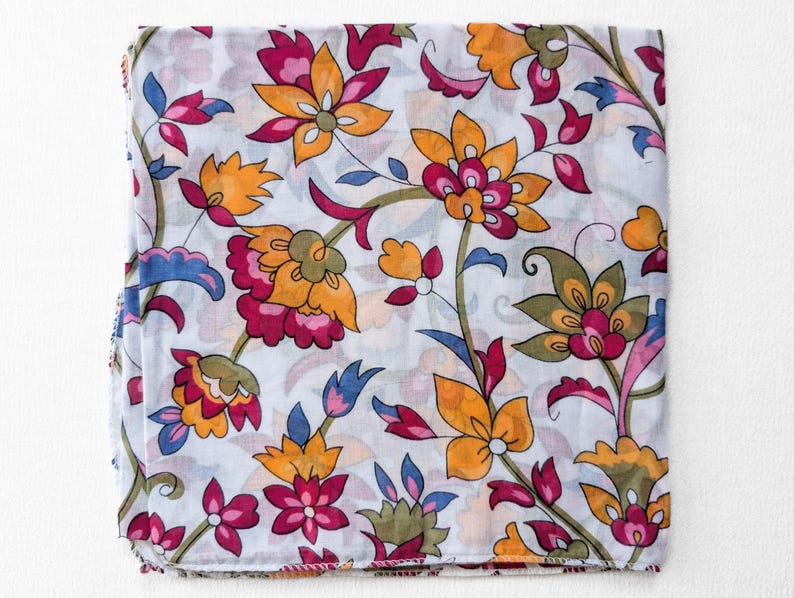 May include: A folded white fabric with a vibrant floral pattern. The design features large, stylized flowers in shades of orange, pink, and green, with blue accents. The fabric is likely a scarf or a similar textile.