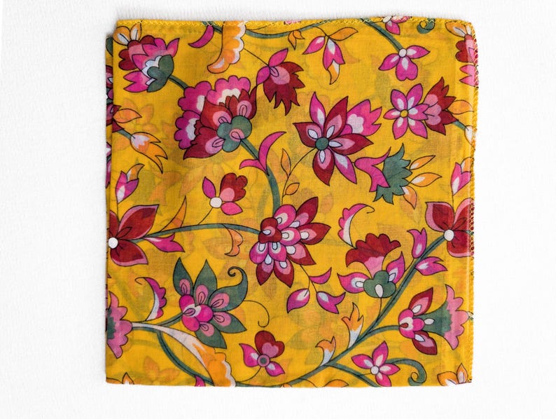 May include: A square, yellow fabric with a floral pattern. The design features pink, red, and green flowers with green leaves and stems. The fabric is folded and appears to be a scarf or handkerchief.