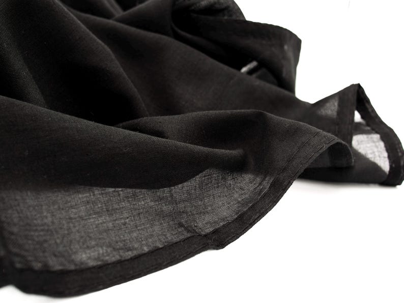 May include: Close-up of a flowing black fabric, possibly linen or a similar lightweight material. The fabric is draped and folded, with visible texture and shadows. The edges are neatly finished.