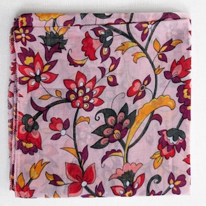May include: A folded, square scarf with a floral pattern. The scarf features a light pink background with a repeating design of flowers in red, purple, yellow, and black. The edges of the scarf are finished with a stitched hem.