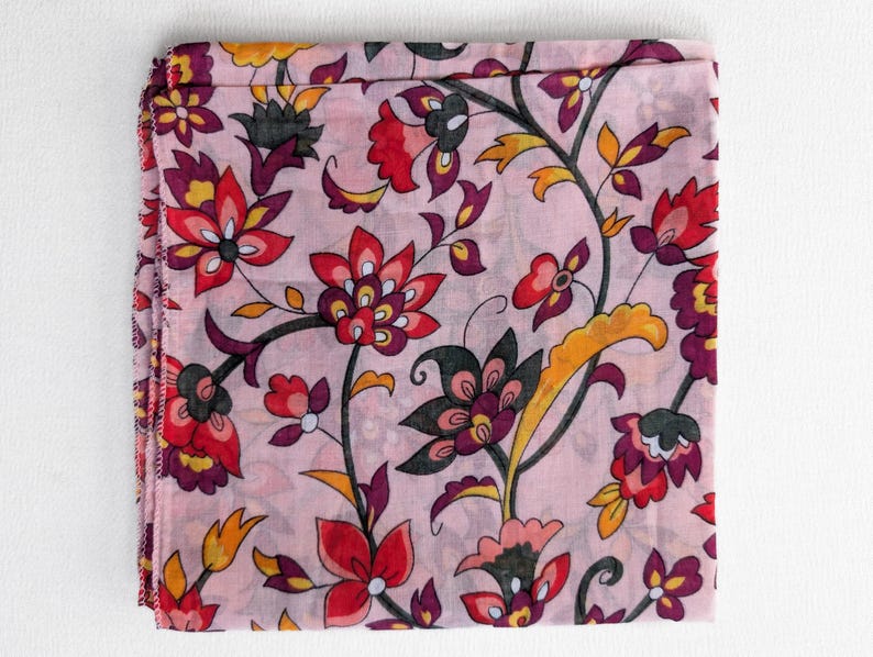 May include: A folded, square scarf with a floral pattern. The scarf features a light pink background with a repeating design of flowers in red, purple, yellow, and black. The edges of the scarf are finished with a stitched hem.
