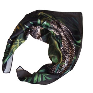 Op de afbeelding: Een zwarte zijden sjaal met een jungledesign. De sjaal heeft een luipaardprint en groene tropische bladeren. De sjaal is opgevouwen en toont de gedetailleerde print en kleurvariaties.